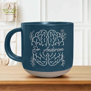May include: A ceramic mug with a dark teal exterior and a light beige base. The mug features a white brain illustration with the name "Dr. Anderson" in script. Floral accents surround the brain design. The mug sits on a wooden surface.