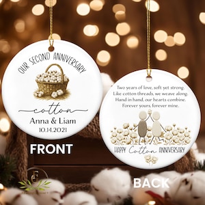 May include: A white ceramic ornament with gold string features a basket of cotton and the text "OUR SECOND ANNIVERSARY." The front says "cotton Anna & Liam 10.14.2021." The back says "HAPPY Cotton ANNIVERSARY" with a couple and cotton.