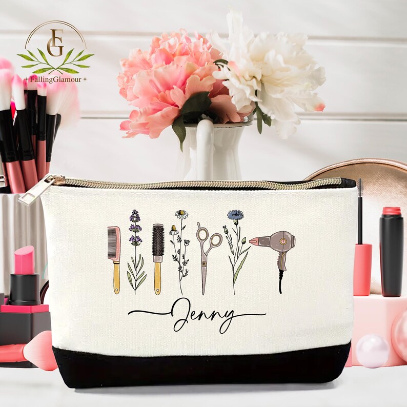 May include: A white cosmetic bag with a black bottom and gold zipper. The bag features illustrations of beauty tools and flowers. The name "Jenny" is written in cursive. Makeup brushes and flowers are in the background.