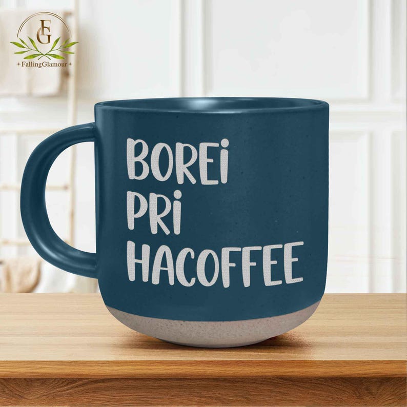 May include: A dark blue ceramic coffee mug with a white base and a large handle. The mug features the words "BOREI PRI HACOFFEE" in white block letters. The mug is sitting on a wooden surface.