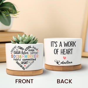 May include: White ceramic planter with a bamboo base. The front of the planter has a heart-shaped design with the words "PATIENT", "CARE", "COOL", "KIND", "CALM", "LISTEN", "GUIDE", "SUPPORT", "SOCIAL WORKER", "HELPFUL", "COMMITTED", "HEART", "ENCOURAGE", and "DEDICATED" written in colourful letters. The back of the planter has the words "IT'S A WORK OF HEART" written in black and the name "Rabaclina" written in black below.