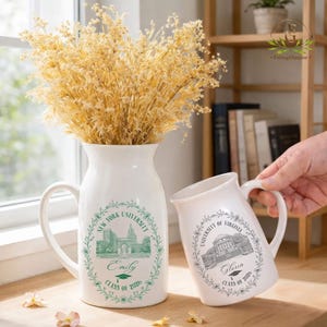 Pu&ograve; includere: Vaso e tazza in ceramica bianca con manici, con emblemi universitari e nomi. Il vaso mostra "New York University" e "Emily, Class of 2026". La tazza mostra "University of Virginia" e "Gloria, Class of 2026". Entrambi con dettagli verdi e grigi.