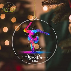 May include: A circular clear acrylic ornament featuring a colorful galaxy-patterned silhouette of a gymnast performing a handstand. The design includes the name Isabella and the year 2026 in white script text, suspended by a gold string.