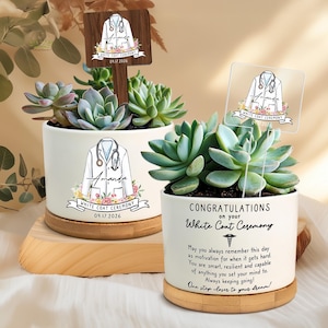 May include: Two white ceramic planters with succulents, personalized for a White Coat Ceremony. Each planter features a medical coat design, the name "Amanda", and the date 09.17.2026. One planter has a wooden sign, the other has a clear acrylic sign.