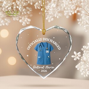 K&ouml;nnte beinhalten: Herzf&ouml;rmiger Glasornament mit blauem Krankenschwester-Uniform-Design. Der Ornament tr&auml;gt die Aufschriften &bdquo;OFFICIALLY DISCHARGED&ldquo; und &bdquo;Retired Nurse 2026&ldquo;. Eine goldene Kette ist zum Aufh&auml;ngen befestigt.