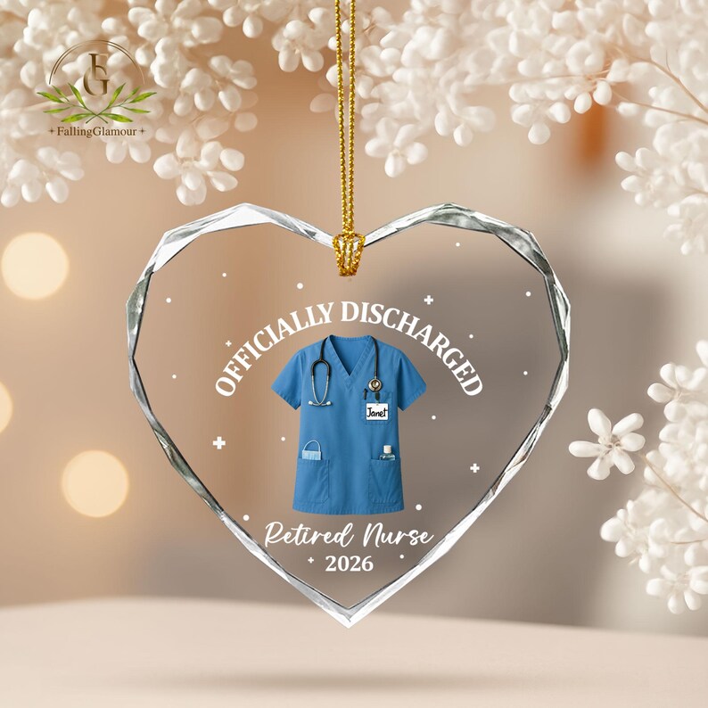 K&ouml;nnte beinhalten: Herzf&ouml;rmiger Glasornament mit blauem Krankenschwester-Uniform-Design. Der Ornament tr&auml;gt die Aufschriften &bdquo;OFFICIALLY DISCHARGED&ldquo; und &bdquo;Retired Nurse 2026&ldquo;. Eine goldene Kette ist zum Aufh&auml;ngen befestigt.