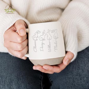 May include: Cream-colored ceramic mug with a gray base, featuring four engraved bunny silhouettes and names: Clover, Daisy, Willow, and Mochi. The mug is held by a person wearing a white sweater and blue jeans.