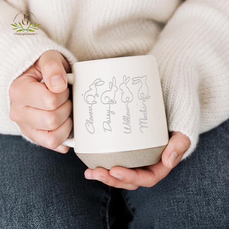 May include: Cream-colored ceramic mug with a gray base, featuring four engraved bunny silhouettes and names: Clover, Daisy, Willow, and Mochi. The mug is held by a person wearing a white sweater and blue jeans.