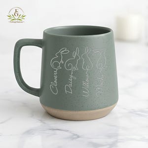May include: A speckled sage green ceramic mug with a light tan base. The mug features four white line-drawn rabbits with names: Clover, Daisy, Willow, and Mochi. The mug has a large handle and is suitable for hot beverages.