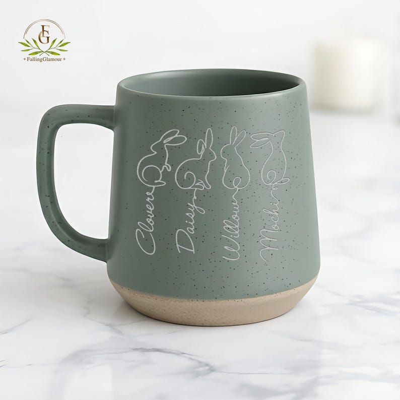 May include: A speckled sage green ceramic mug with a light tan base. The mug features four white line-drawn rabbits with names: Clover, Daisy, Willow, and Mochi. The mug has a large handle and is suitable for hot beverages.
