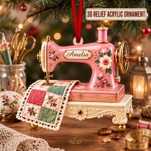 May include: A pink 3D relief acrylic ornament shaped like a vintage sewing machine with gold accents and floral details. The machine features the name Amelia in script, a spool of pink thread, and a miniature quilted fabric piece with lace trim.