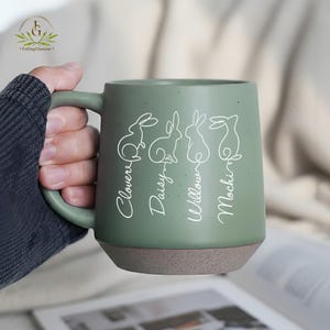 May include: A sage green ceramic mug with a brown base, featuring white line drawings of rabbits and the names "Clover", "Daisy", "Willow", and "Mochi". The mug has a large handle and a speckled finish.