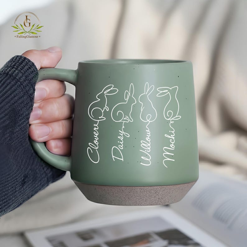 May include: A sage green ceramic mug with a brown base, featuring white line drawings of rabbits and the names "Clover", "Daisy", "Willow", and "Mochi". The mug has a large handle and a speckled finish.