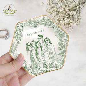 May include: A white ceramic trinket dish with a gold rim and a green floral design. The dish features a sketch of a group of people and the text "Bestfriends for life". The dish is held in a hand.