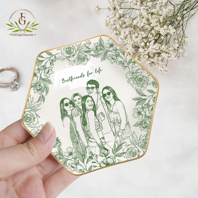 May include: A white ceramic trinket dish with a gold rim and a green floral design. The dish features a sketch of a group of people and the text "Bestfriends for life". The dish is held in a hand.