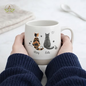 May include: Cream-coloured ceramic mug with illustrations of two cats, a tortoiseshell and a grey one, with the names "Miny" and "Bella" beneath. The mug has a handle and is decorated with paw prints, a heart, and a ball of wool.
