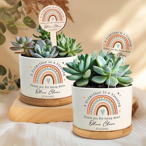 May include: Two white ceramic planters with succulents, each featuring a rainbow design and the text "A Godmother is a blessing. Thank you for being mine. Olivia Claire 07.12.2025." One planter sits on a wooden base.