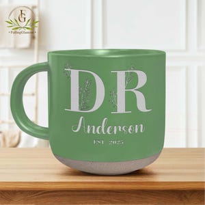 May include: Green ceramic mug with a white inscription that reads "DR Anderson EST. 2025". The mug has a white floral design around the letters.