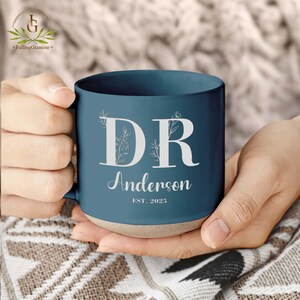 May include: A blue ceramic mug with a white design featuring the initials "DR" with floral accents, the name "Anderson", and the text "EST. 2025".