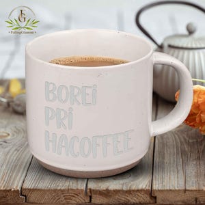 May include: A white ceramic mug filled with coffee, with the words "BOREI PRI HACOFFEE" in light grey. The mug has a brown base and a large handle. The mug is on a wooden surface.