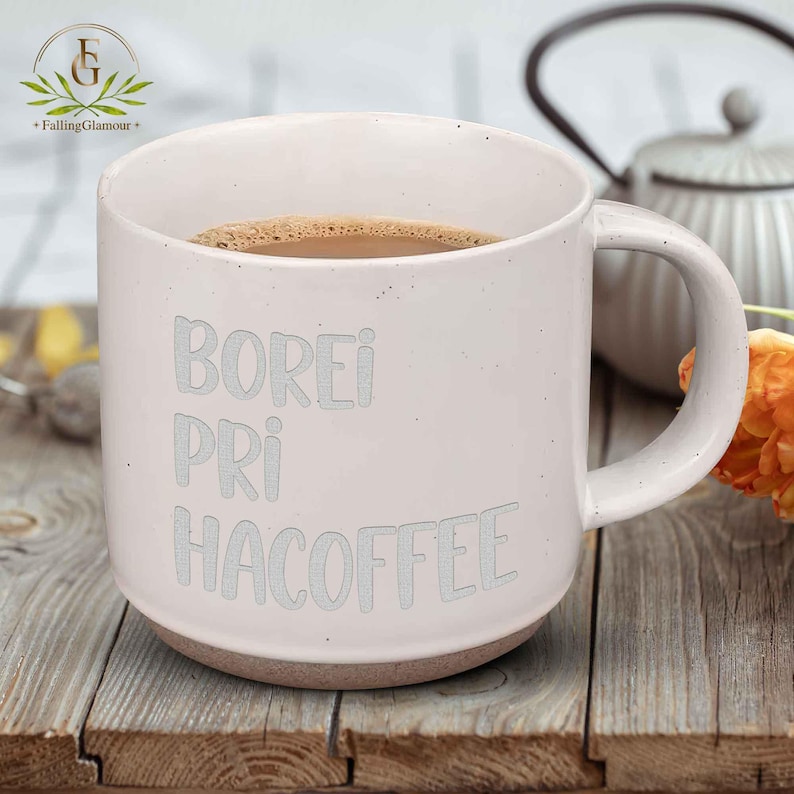 May include: A white ceramic mug filled with coffee, with the words "BOREI PRI HACOFFEE" in light grey. The mug has a brown base and a large handle. The mug is on a wooden surface.