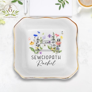 May include: A white, square trinket dish with a gold rim. The dish features a sewing machine illustration surrounded by colourful flowers and butterflies. The text "SEWCIOPATH Rachel" is printed below the illustration.