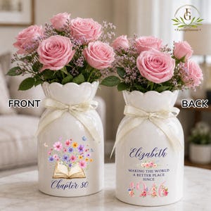 May include: Two white ceramic vases with scalloped rims and cream ribbons, each holding pink roses and purple filler flowers. The front vase features an open book graphic with flowers and the text Chapter 80. The back vase displays the name Elizabeth, the text Making the world a better place since 1946, and floral accents.