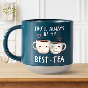 May include: A teal blue ceramic mug with a white rim and base. The mug features a graphic of two cartoon teacups with smiling faces and the text "You'll always be my best-tea". The teacups are labelled "Annie" and "Sophia".