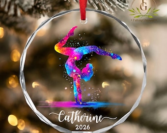 May include: A circular clear glass ornament with faceted edges, featuring a colorful galaxy-patterned gymnast performing a handstand. The name Catherine and the year 2026 are printed in white script and block text. A red ribbon is attached to the top.