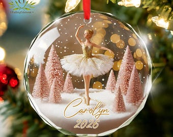 May include: A clear, faceted, circular glass ornament hanging from a red ribbon. Inside, a ballerina in a gold and white tutu stands amidst pink bottle brush trees and white snow. The text "Carolyn" and "2026" is printed in gold script.