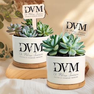 May include: Two white ceramic plant pots with succulents, each on a wooden base. The pots have the text "DVM Dr. Melissa Jamison DOCTOR OF VETERINARY MEDICINE" in black. One pot has a wooden sign with the same text.