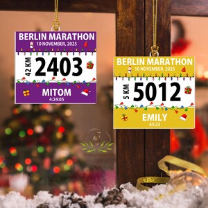May include: Two square hanging ornaments featuring Berlin Marathon race bib designs. One is purple with the number 2403 and name MITOM, the other is yellow with the number 5012 and name EMILY. Both include the date 10 November 2025 and holiday graphics.