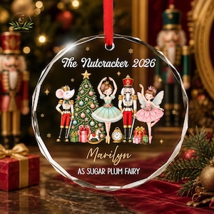 May include: A round, faceted glass Christmas ornament hanging from a red ribbon. The design features a Christmas tree, a mouse, a ballerina, a nutcracker, and a fairy with the text "The Nutcracker 2026", "Marilyn", and "AS SUGAR PLUM FAIRY".