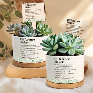 May include: Two white ceramic pots with succulents, each on a wooden base. The pots have the text "Difference Maker" and a definition. A wooden sign and a clear sign with the same text are placed in the pots.