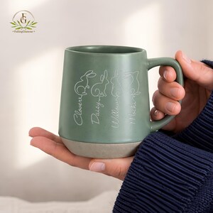 May include: Green ceramic mug with a light beige base. The mug features white line drawings of rabbits and the names "Clover", "Daisy", "Willow", and "Mochi" in a script font. The mug has a large handle.