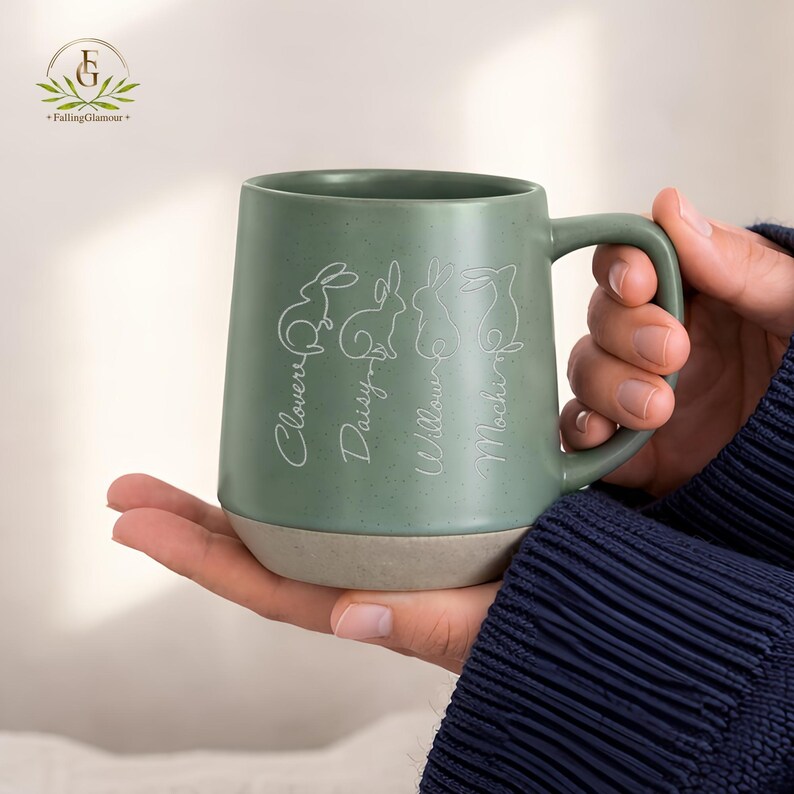 May include: Green ceramic mug with a light beige base. The mug features white line drawings of rabbits and the names "Clover", "Daisy", "Willow", and "Mochi" in a script font. The mug has a large handle.