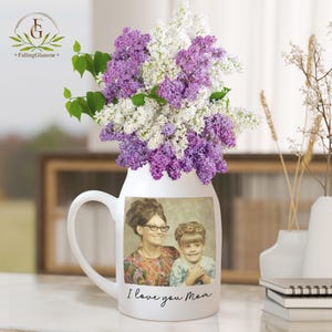 May include: White ceramic pitcher with a handle, featuring a photo of two women and the text "I love you Mom". The pitcher is filled with a bouquet of purple and white lilac flowers.