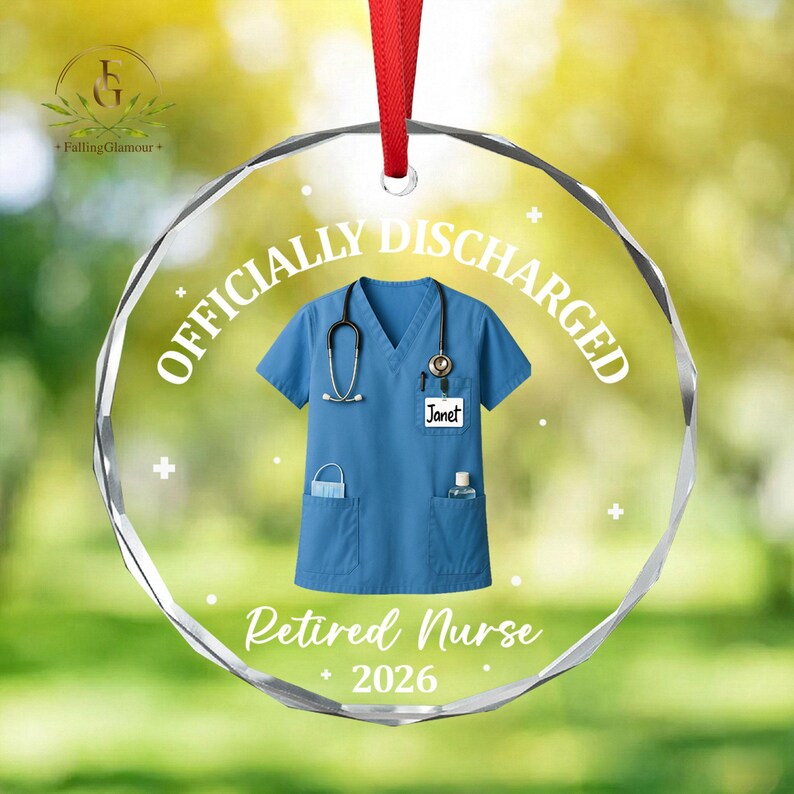 K&ouml;nnte beinhalten: Ein klarer, runder Weihnachtsschmuck mit rotem Band. Der Schmuck zeigt eine blaue Krankenschwesteruniform mit Stethoskop, den Namen "Janet" und die Texte "OFFICIALLY DISCHARGED" und "Retired Nurse 2026".