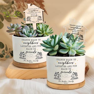 May include: Two white ceramic planters with succulents, each on a wooden base. The planters have the text "CHANCE MADE US neighbours, LAUGHTER AND FUN MADE US friends" with house illustrations. Wooden house-shaped signs are in the planters.