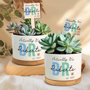 May include: Two white ceramic planters with succulents, each featuring the text "Actually It's Roberts" and "est. 2025". One planter sits on a wooden base, the other on a bamboo coaster. The planters have a blue and green colour scheme.