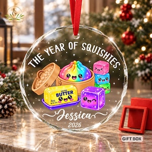 May include: A round clear glass ornament with a red ribbon hanger featuring the text THE YEAR OF SQUISHIES, Jessica, and 2026. The graphic shows a rainbow dumpling in a bamboo steamer, a stick of salted butter labelled 4oz, and three colourful cubes.