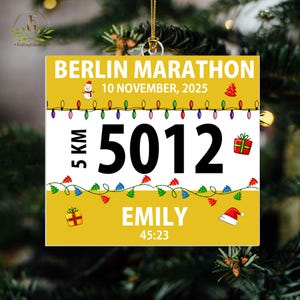 May include: A square yellow and white ornament featuring the text BERLIN MARATHON, 10 NOVEMBER, 2025, 5 KM, 5012, EMILY, and 45:23. The design includes colorful Christmas light strings, a snowman, a gift box, and a Santa hat on a gold string.