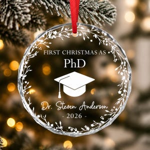 May include: A circular clear glass ornament with faceted edges, featuring a white printed wreath design. The text reads FIRST CHRISTMAS AS PhD, Dr. Steven Anderson, and 2026, with a central graduation cap icon. A red ribbon is attached for hanging.