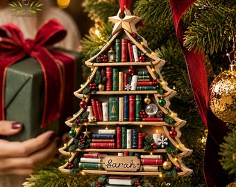 May include: A Christmas tree-shaped wooden ornament featuring multiple shelves filled with miniature books in red, green, and white. The ornament is topped with a gold star, decorated with tiny lights, and includes a nameplate with the name Sarah.