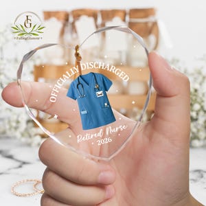 K&ouml;nnte beinhalten: Herzf&ouml;rmiger Glasornament mit blauem medizinischem Kittel-Design, Stethoskop und den Worten "Officially Discharged" und "Retired Nurse 2026". Der Ornament wird an einer Schnur aufgeh&auml;ngt.
