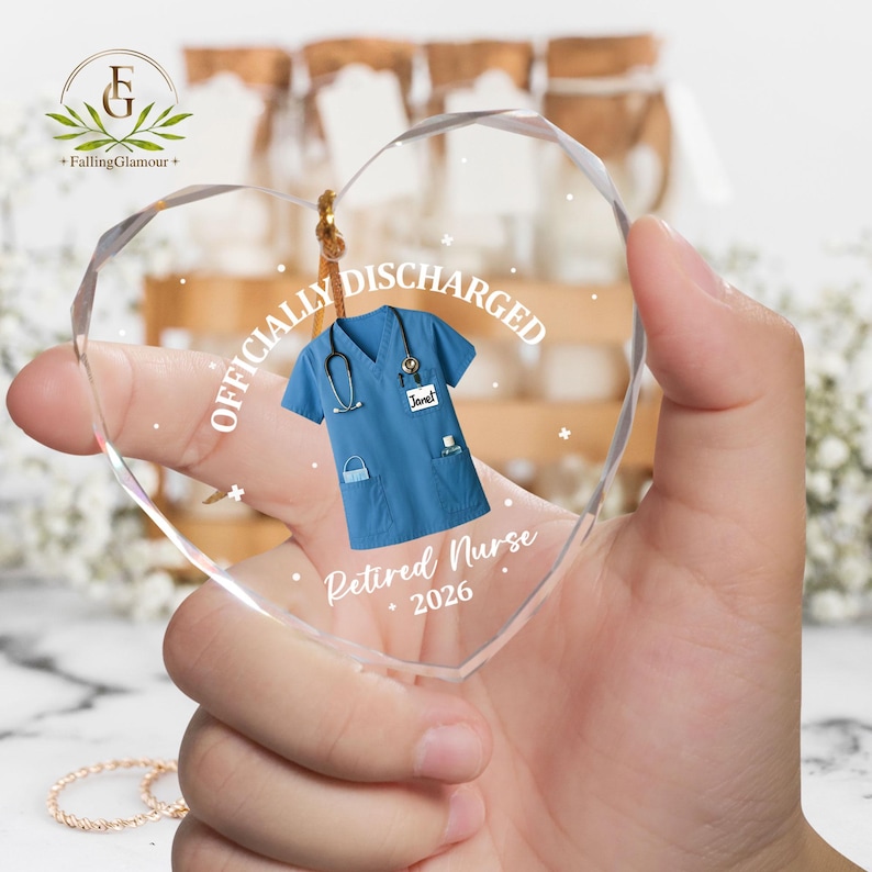 K&ouml;nnte beinhalten: Herzf&ouml;rmiger Glasornament mit blauem medizinischem Kittel-Design, Stethoskop und den Worten "Officially Discharged" und "Retired Nurse 2026". Der Ornament wird an einer Schnur aufgeh&auml;ngt.