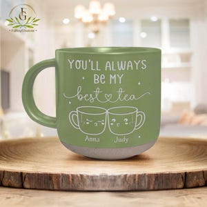 May include: A green ceramic mug with the text "YOU'LL ALWAYS BE MY best tea" and two cartoon tea cups. The mug has a light brown base and a curved handle. The names "Anna" and "Judy" are below the tea cups.