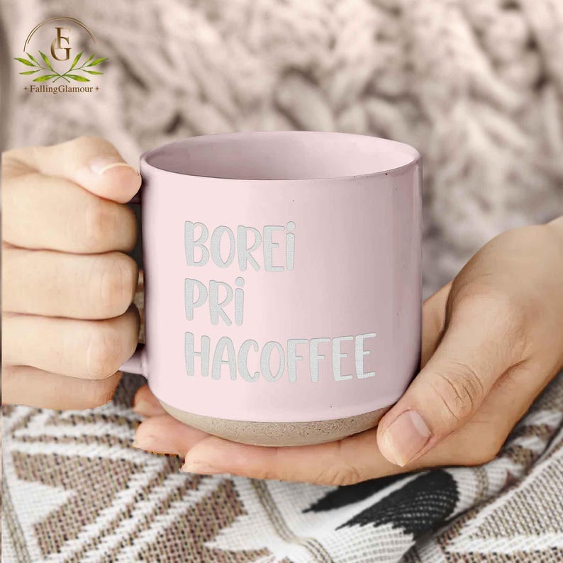 May include: A pale pink ceramic coffee mug with the words "BOREI PRI HACOFFEE" in white lettering. The mug has a light brown base and is held by a person.