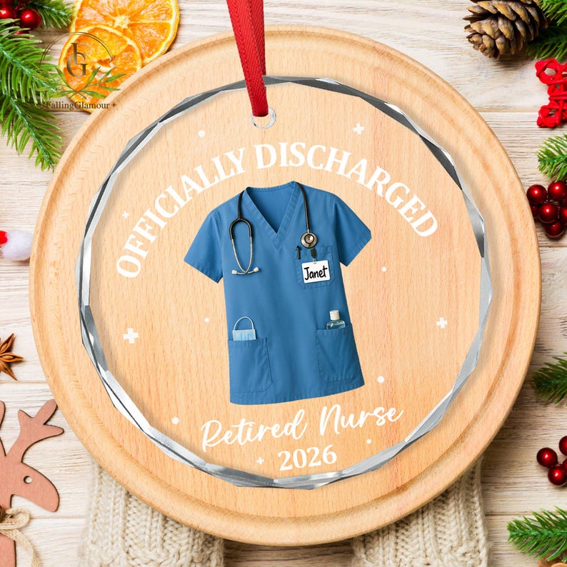 K&ouml;nnte beinhalten: Ein klarer, runder Weihnachtsschmuck mit rotem Band. Der Schmuck zeigt eine blaue Krankenschwesteruniform mit Stethoskop und dem Text "OFFICIALLY DISCHARGED" und "Retired Nurse 2026". Der Name "Janet" steht auf der Uniform.