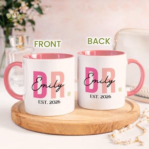May include: Two white ceramic mugs with pink handles and interiors. The mugs feature the text "DR. Emily EST. 2026" in pink and black lettering. The mugs are displayed on a wooden tray.